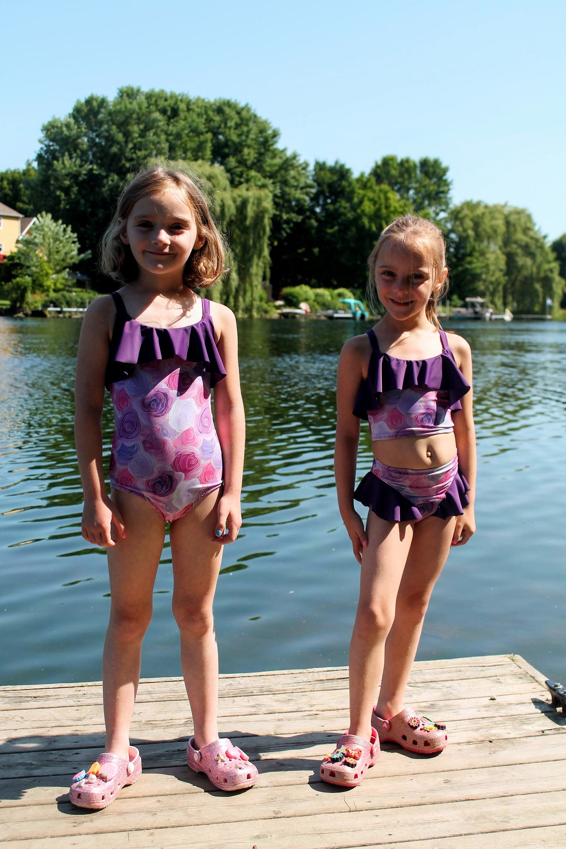 Two young girls in matching swimsuits standing on a dock by a lake.
