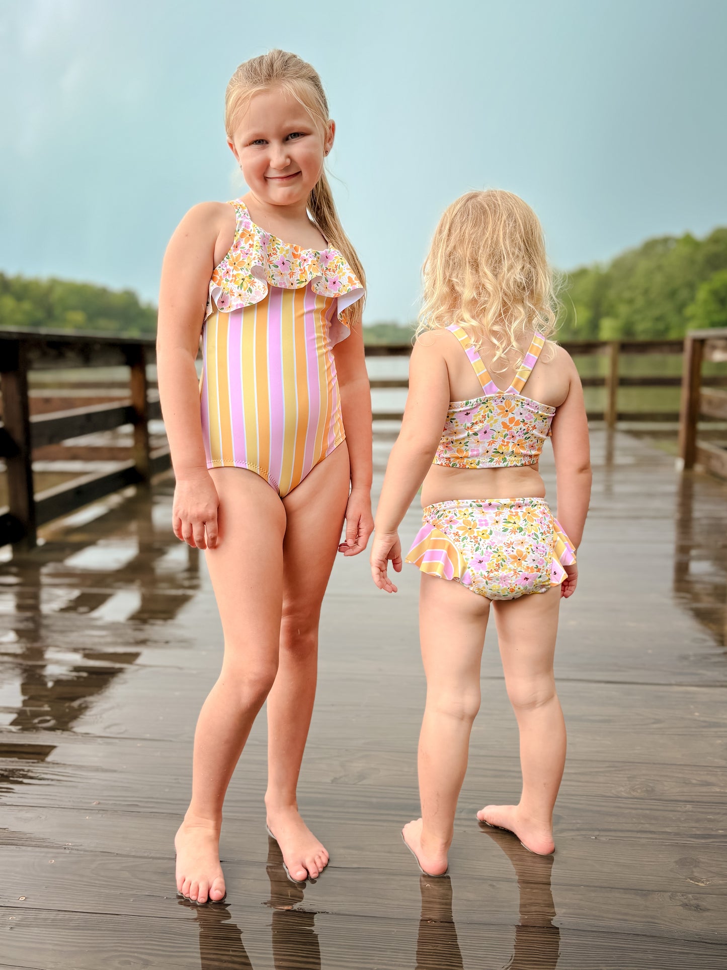 Two young girls in colorful swimsuits standing on a wooden dock.