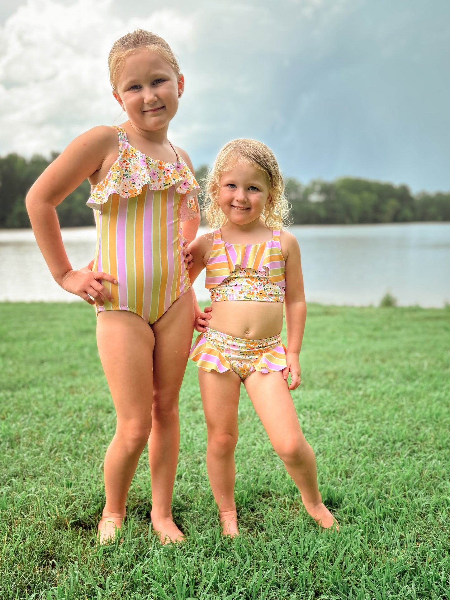 Two young girls in matching swimsuits standing on grass near a body of water.