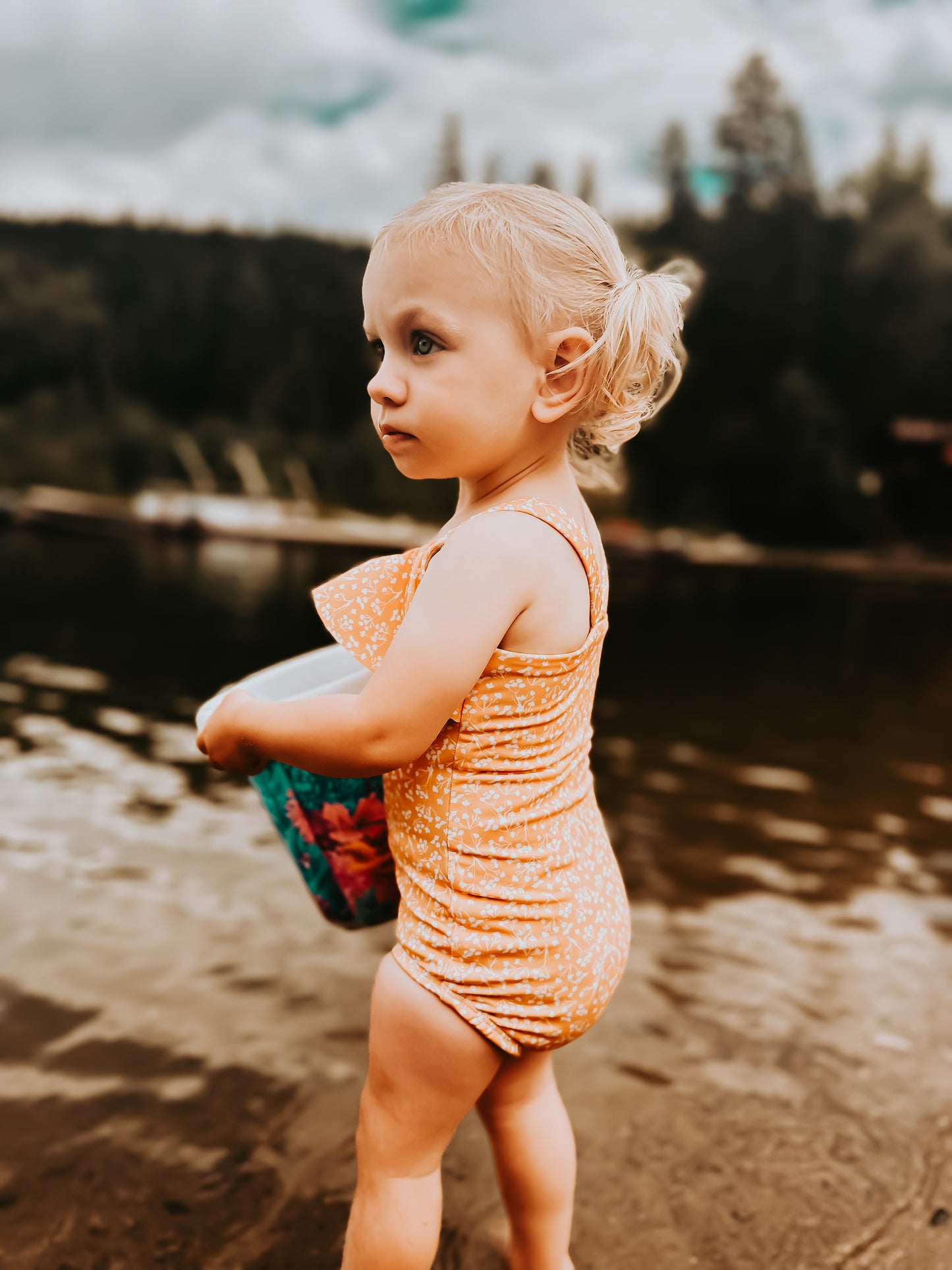 Child in a swimsuit standing by a body of water with trees in the background