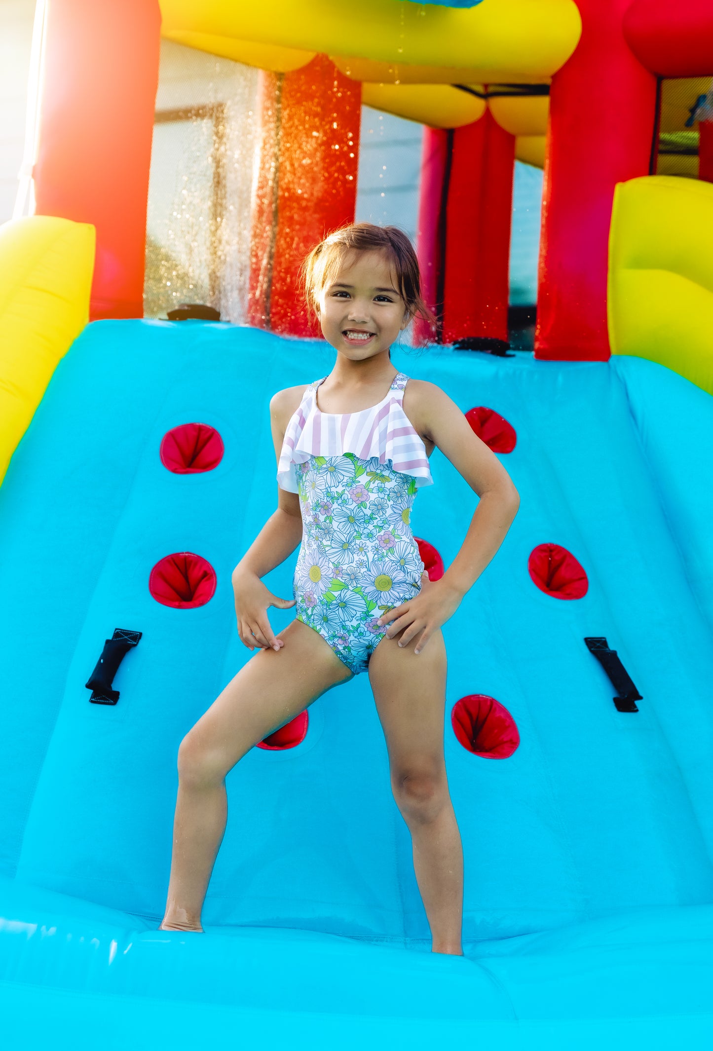 Young girl in a colorful swimsuit standing on a blue inflatable slide with red and black decorations.