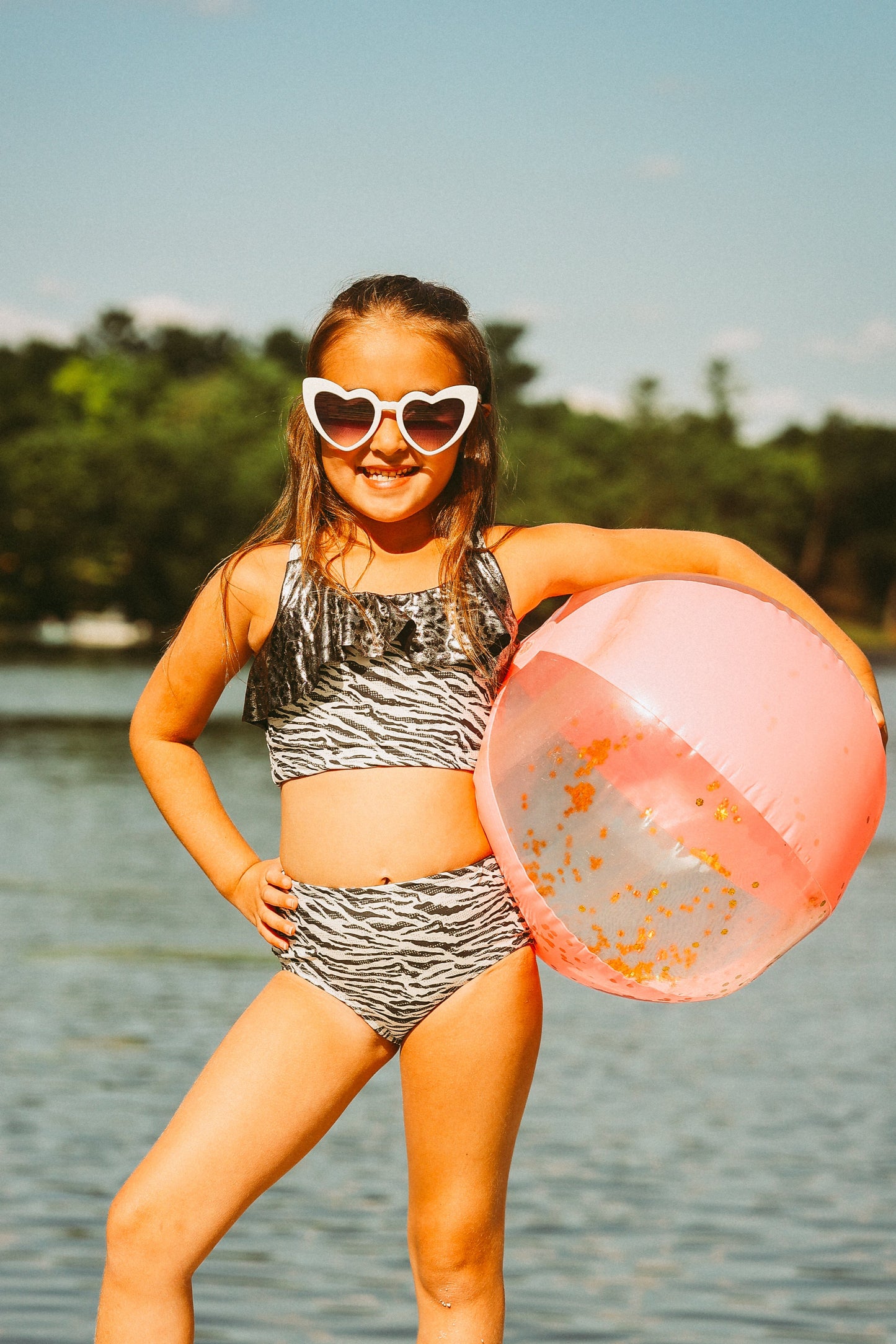 Young girl in a bikini and sunglasses holding a pink beach ball by a lake.