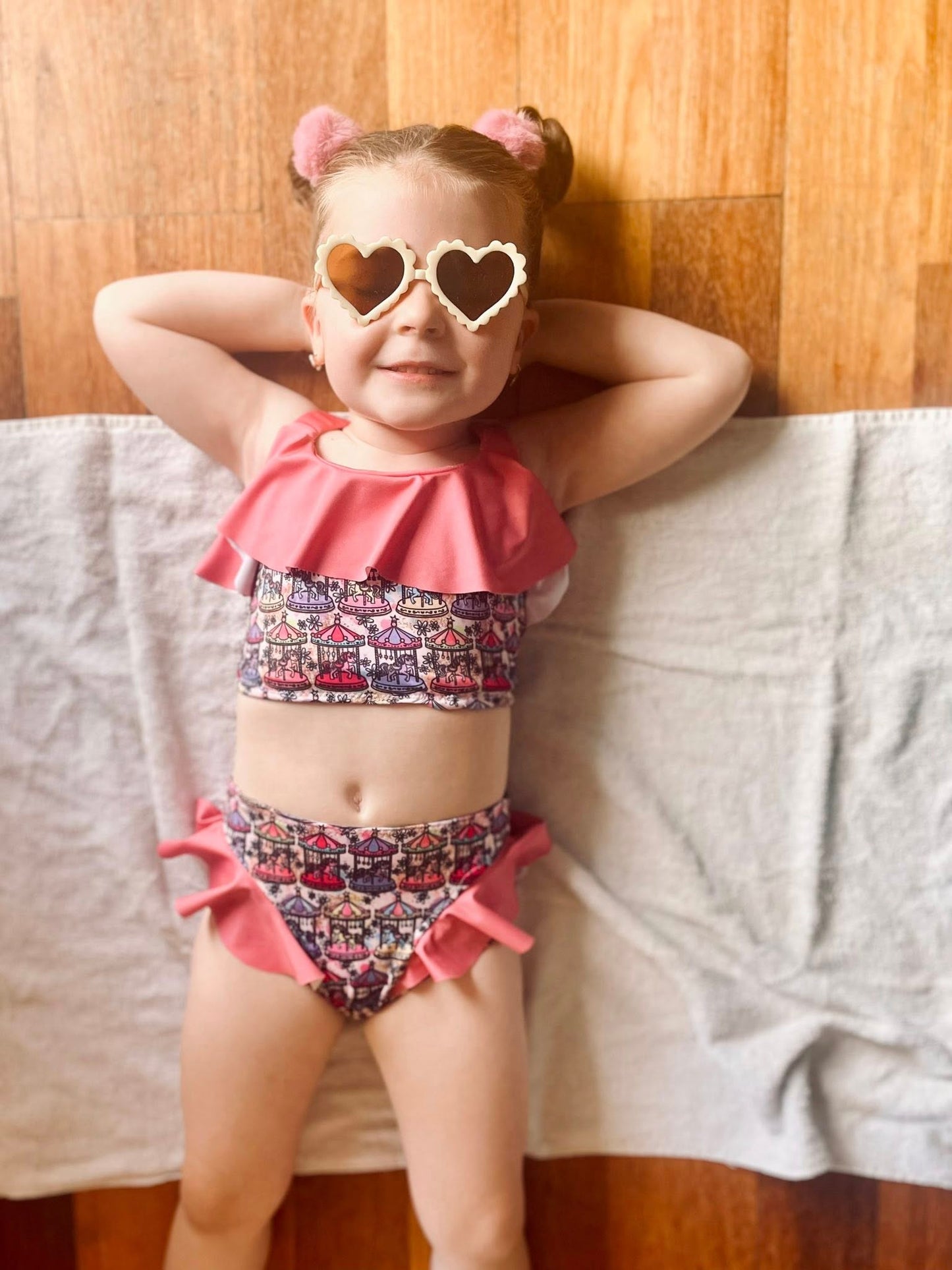 Child wearing a pink and patterned two-piece swimsuit with heart-shaped sunglasses against a wooden wall.