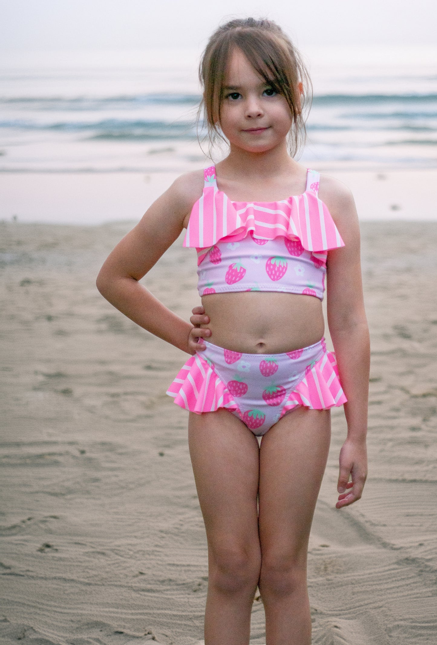 Young girl in a pink and white swimsuit with strawberry pattern on a beach.
