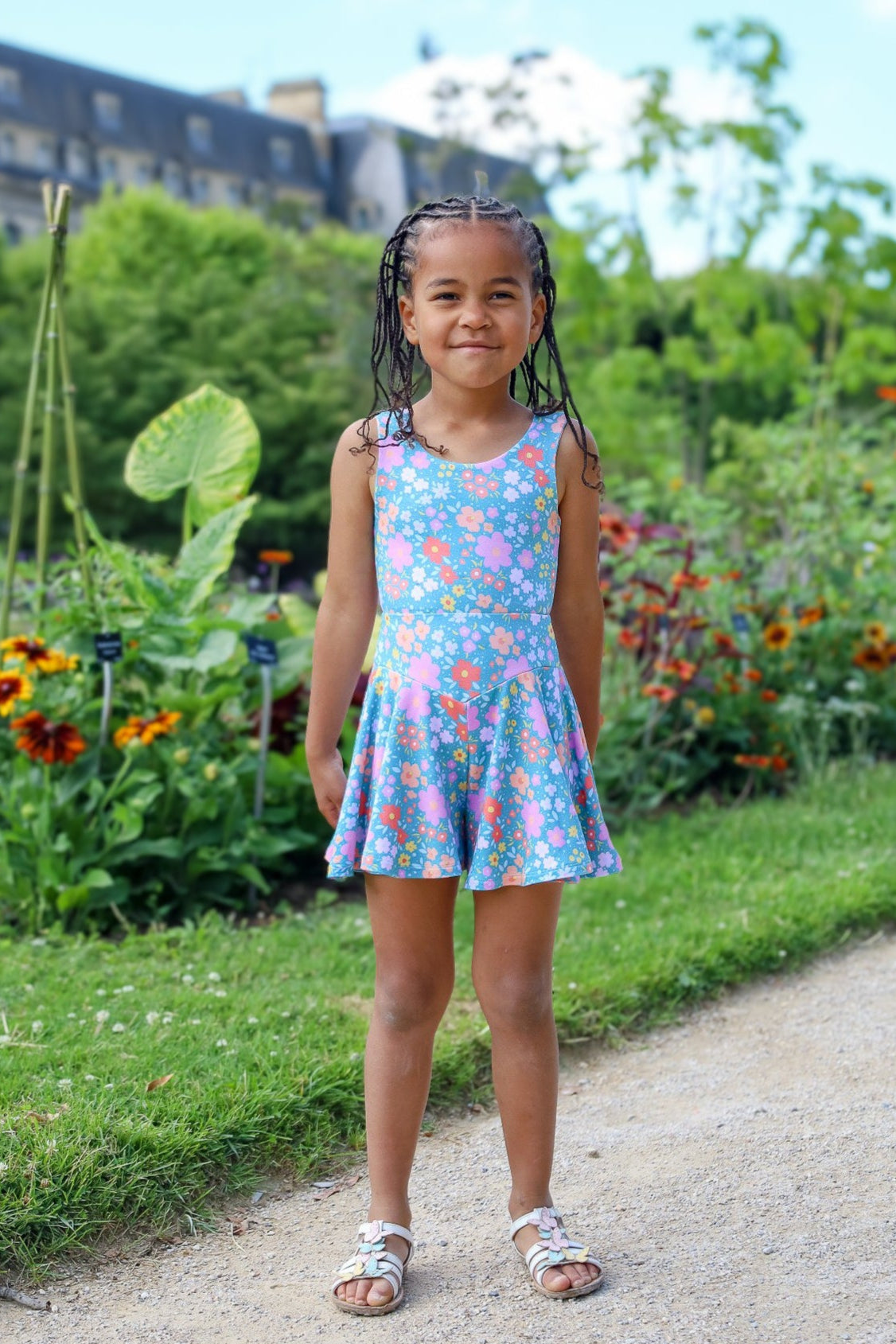 Young girl in a floral dress standing on a path in a garden with colorful flowers and greenery.