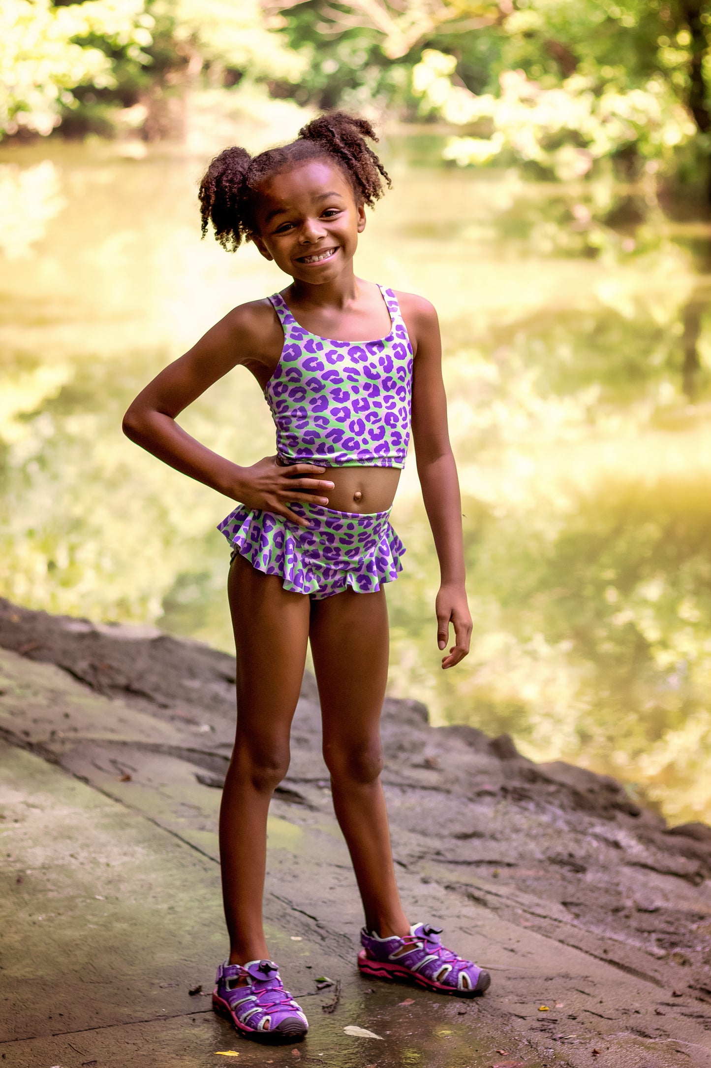 Young girl in a purple swimsuit standing on a log by a body of water with greenery.