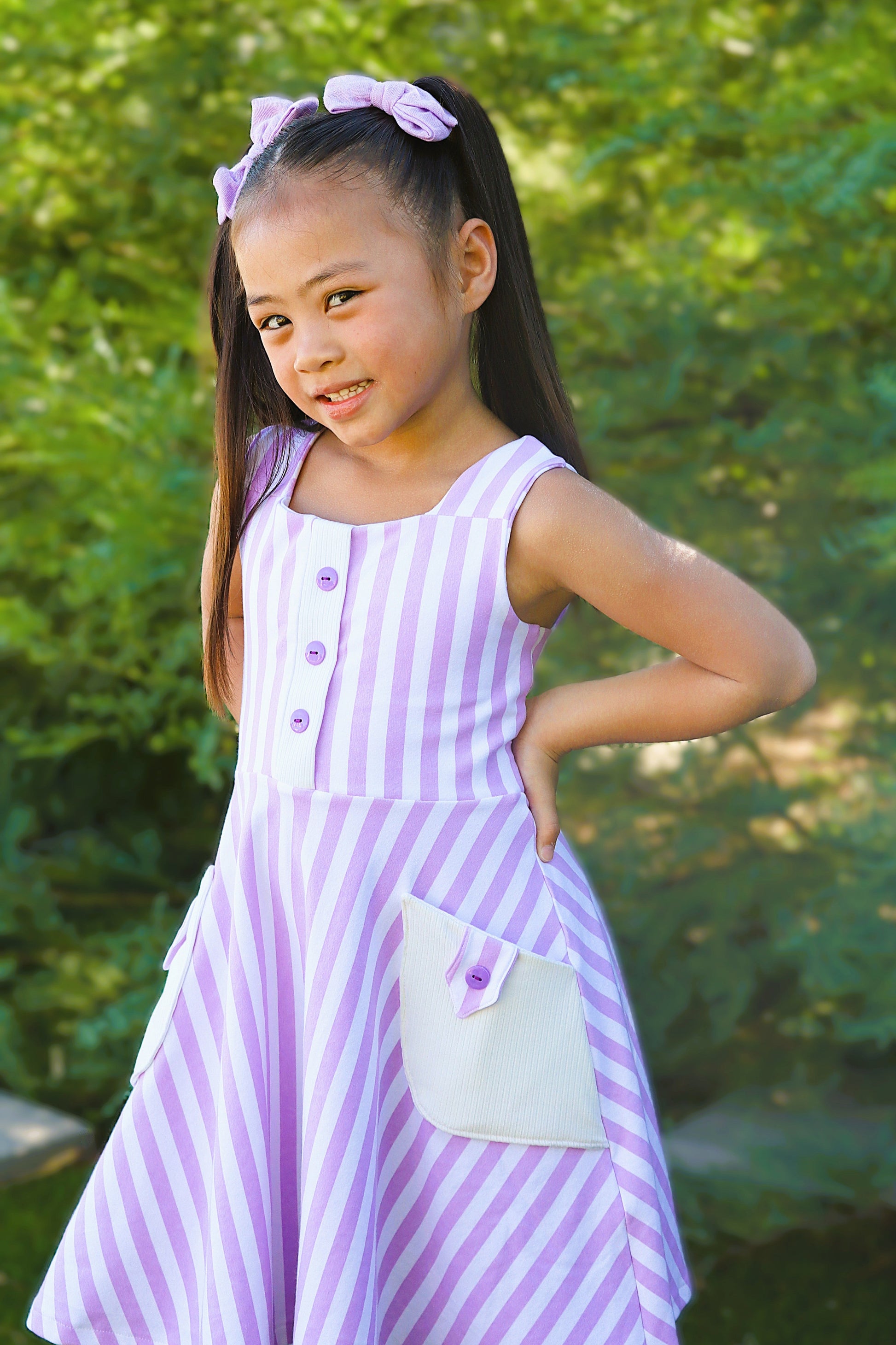 Young girl wearing a purple striped dress with a bow in her hair, standing outdoors.