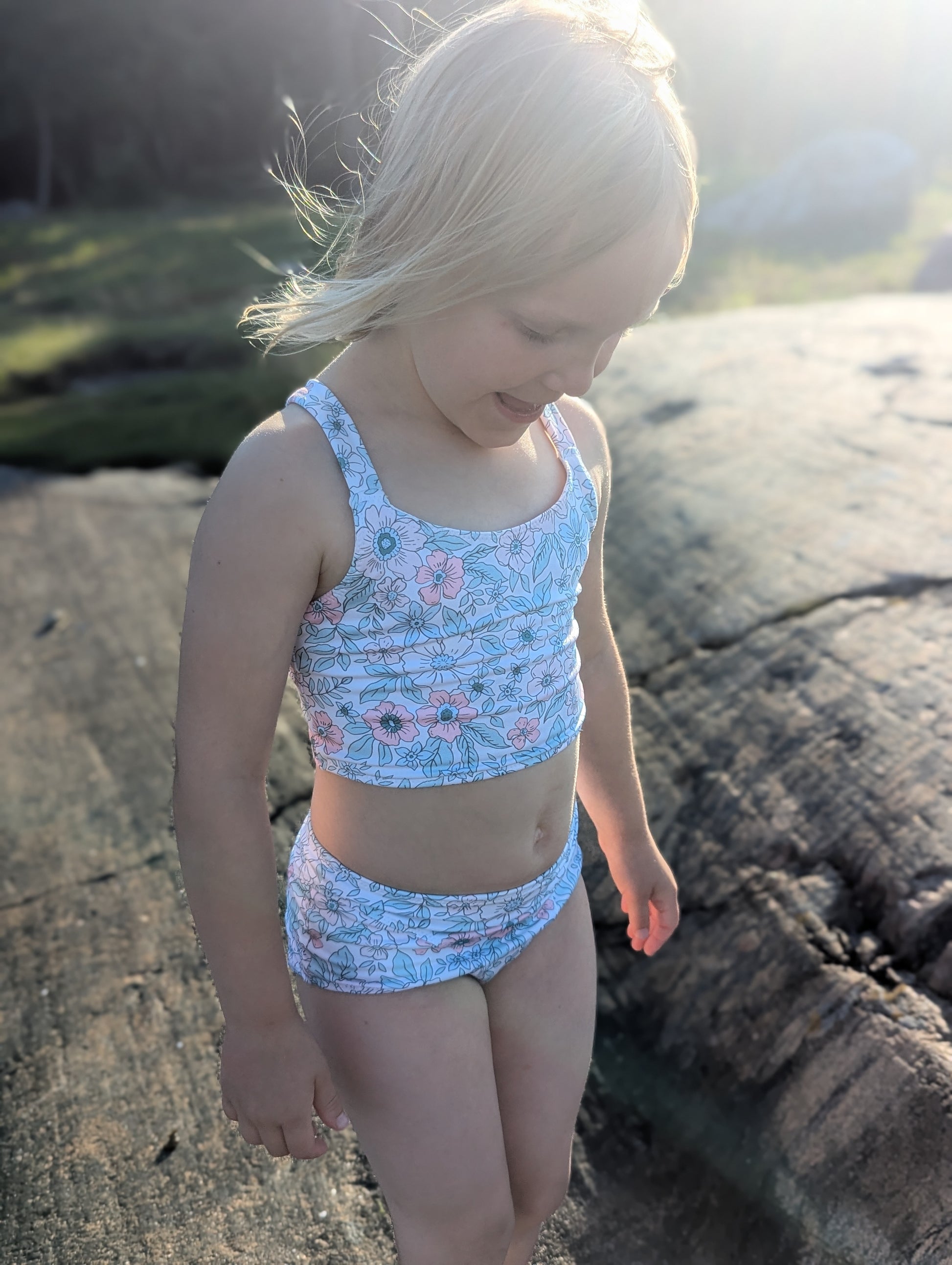 Young girl in a blue floral swimsuit standing on rocks by the water.