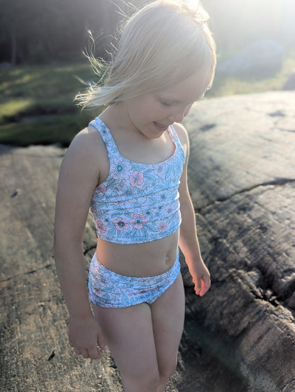 Young girl in a blue floral swimsuit standing on rocks by the water.