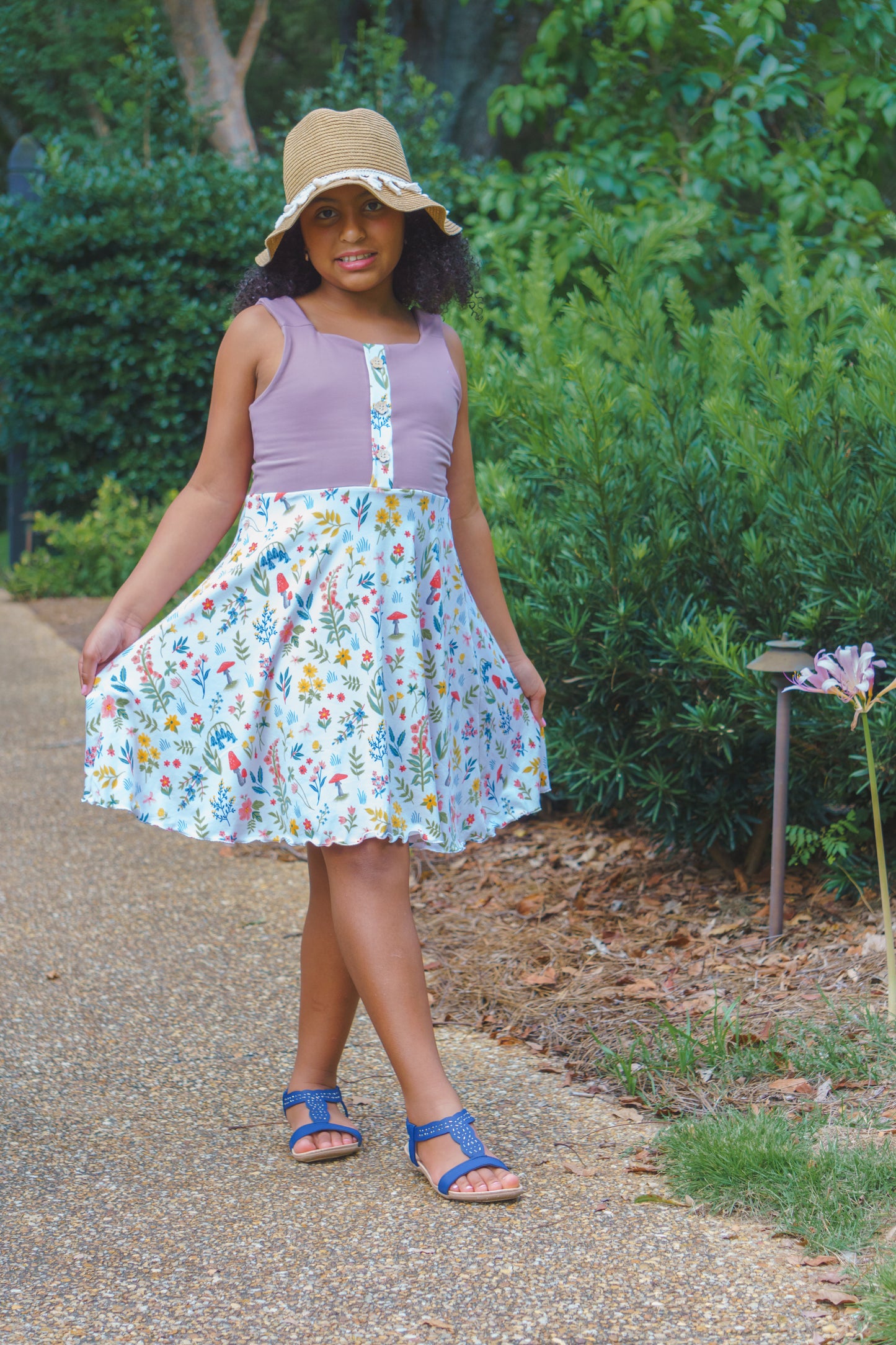 Young girl wearing a floral dress and sun hat standing on a path with greenery in the background