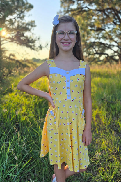 Young girl in a yellow dress standing in a grassy field with trees in the background.
