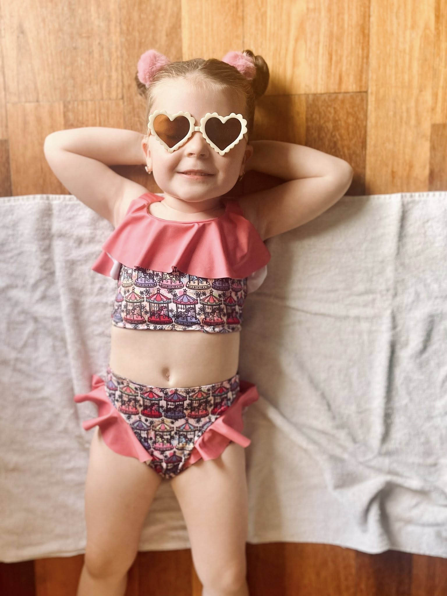 Child wearing a pink and patterned two-piece swimsuit with heart-shaped sunglasses against a wooden wall.