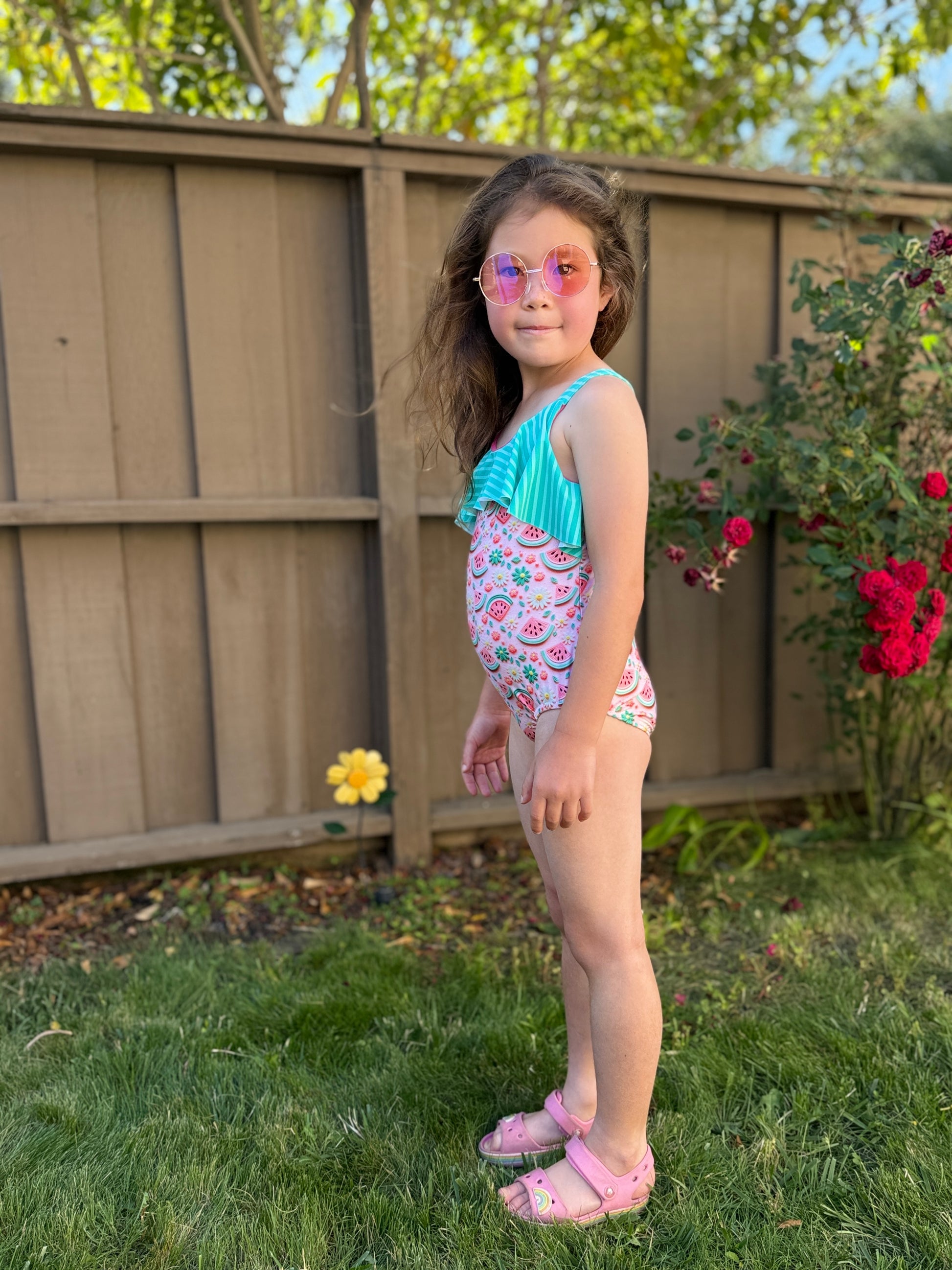Young girl in a colorful swimsuit standing outdoors with a wooden fence and flowers in the background