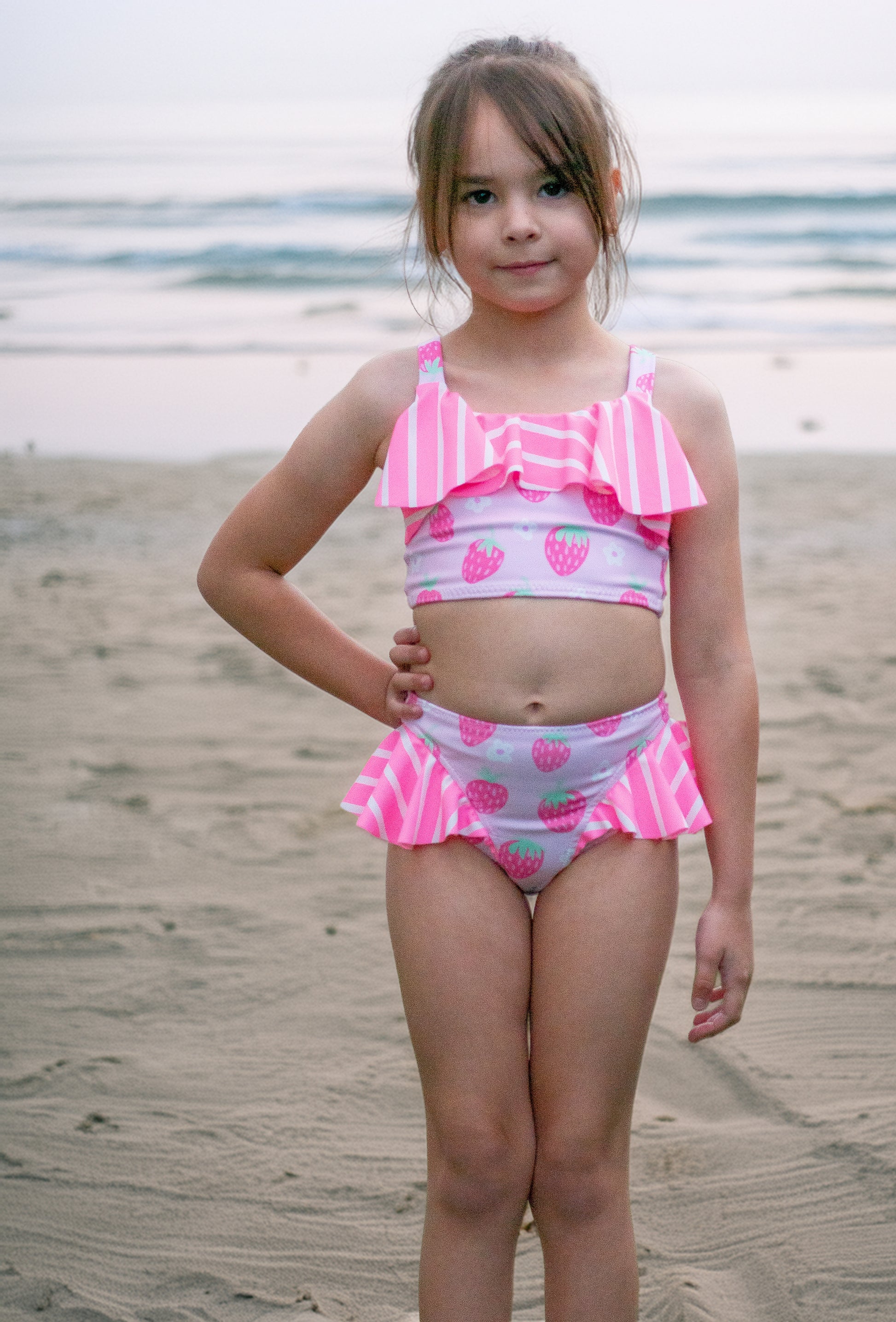 Young girl in a pink and white swimsuit with strawberry pattern on a beach.