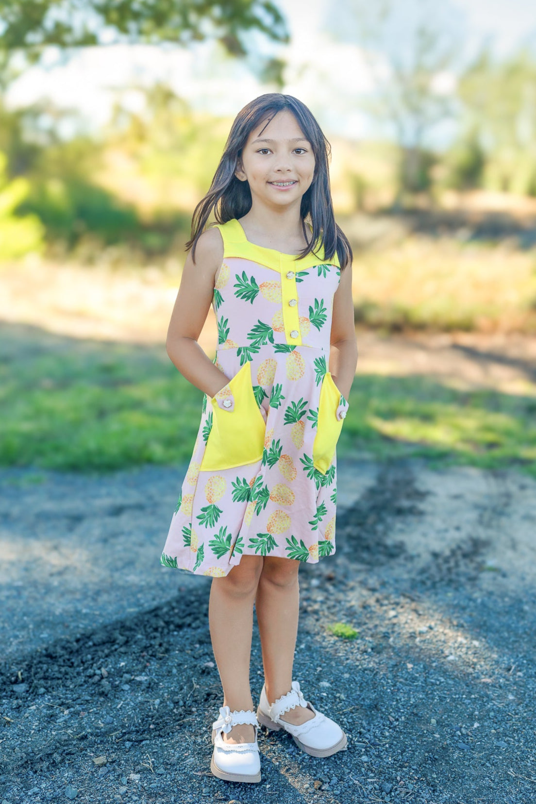 Young girl in a yellow dress with floral pattern standing on a dirt path with greenery in the background