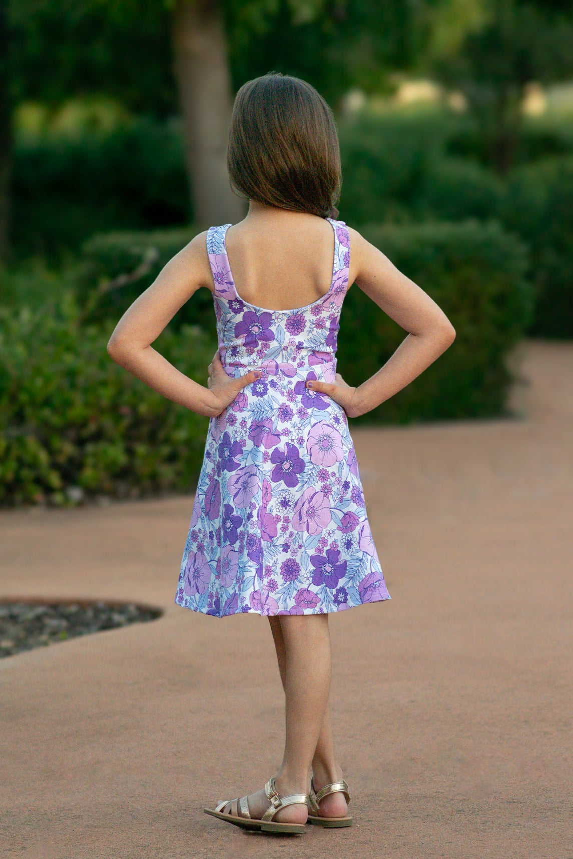 Girl wearing a purple floral dress standing on a path with greenery in the background