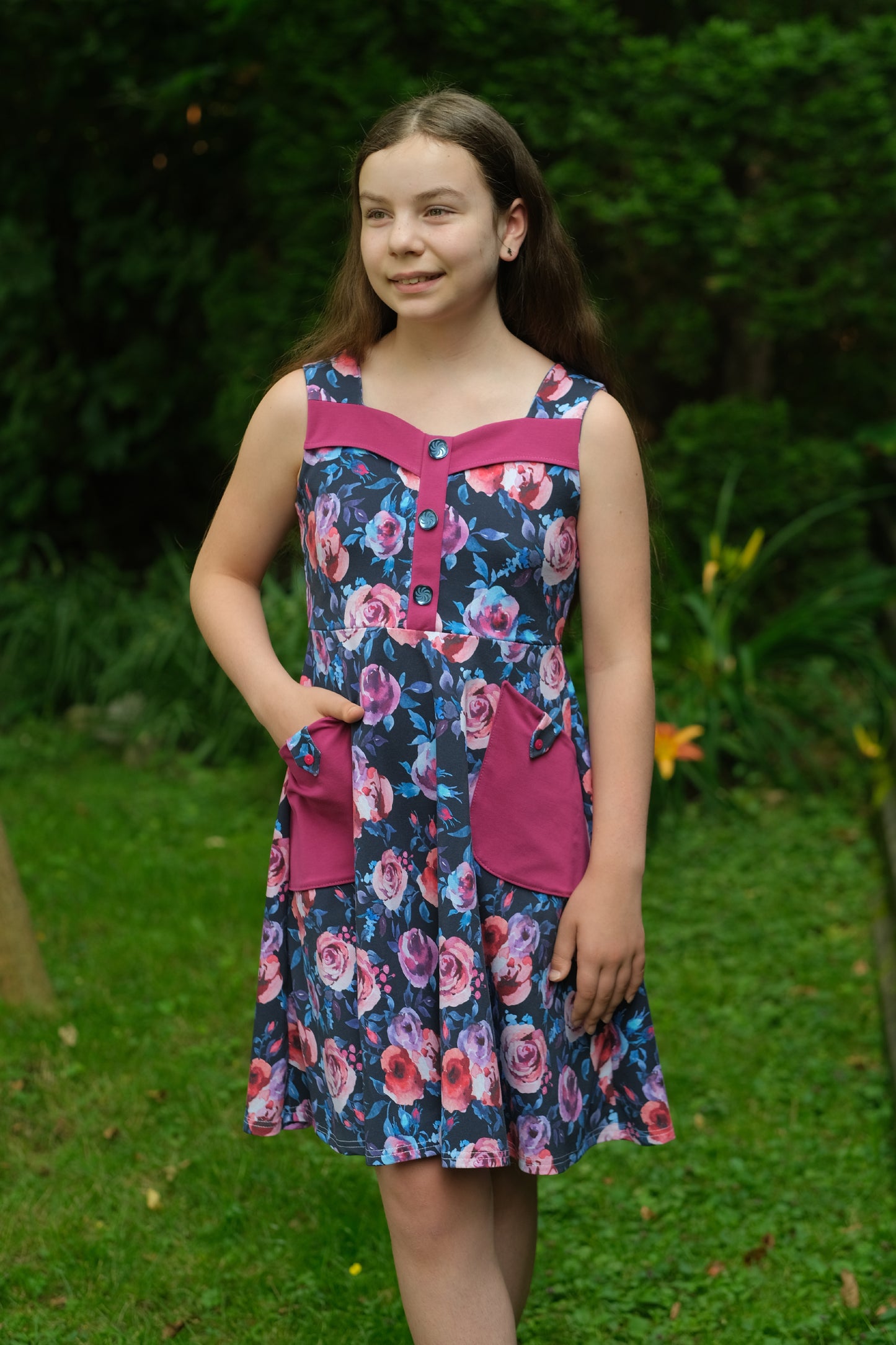 Young girl wearing a floral dress with pink accents standing outdoors on grass.